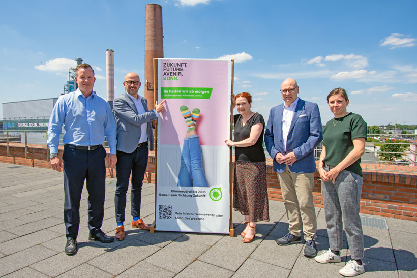 Auf dem Foto sind 5 Personen zu sehen. Die Personen sind SWB-Chef Olaf Hermes, BonnNetz-Geschäftsführer Urs Reitis, Oberbürgermeisterin Katja Dörner, Beigeordneter Helmut Wiesner und Dr. Giulia Pugnaghi. Sie stehen auf einem Dach, im Hintergrund zu sehen ist das Heizkraftwerk Nord. Zwischen ihnen steht ein mannshohes Plakat mit dem Slogan "So heizen wir morgen".